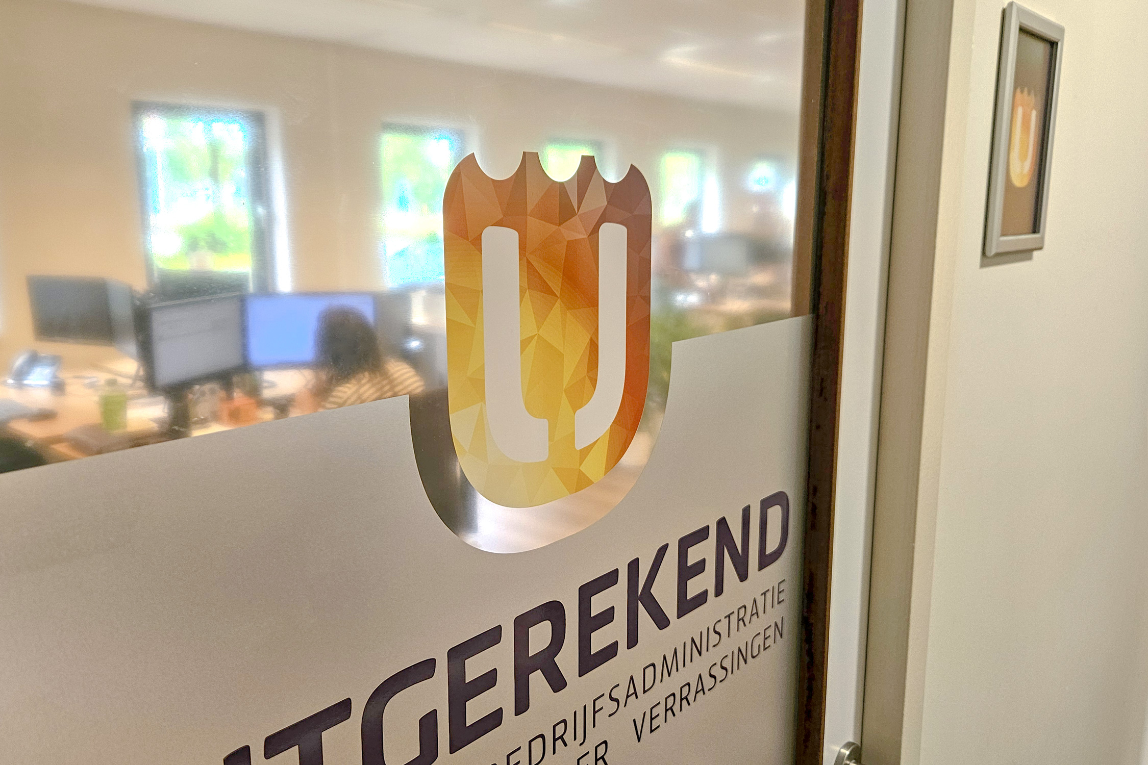 Privacyfolie binnendeur Uitgerekend Hier Close-up van de privacyfolie op de binnendeur van Uitgerekend Hier, met het beeldmerk en een vaag zicht op het kantoor erachter