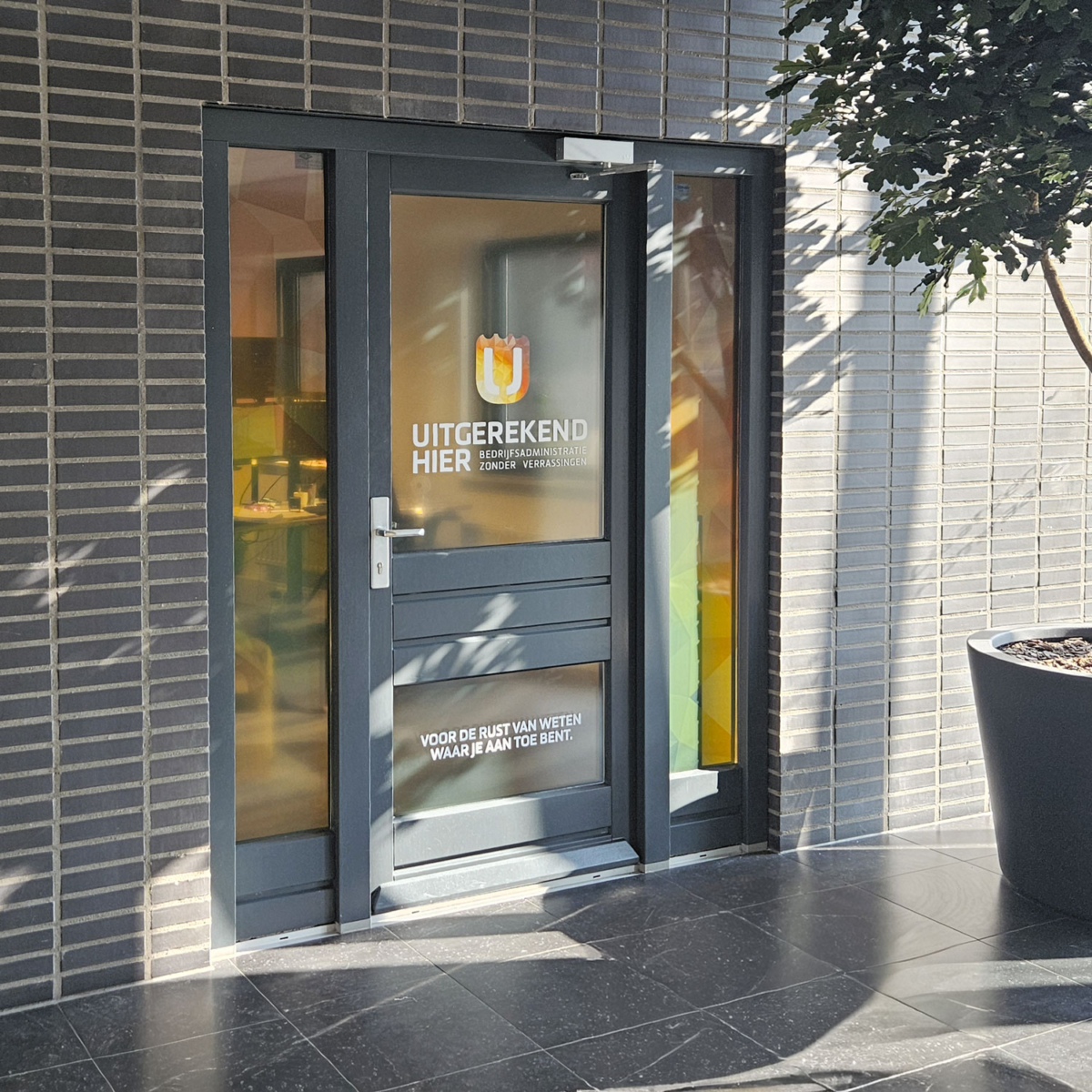 Entree Uitgerekend Hier met raambelettering Entreedeur van Uitgerekend Hier met logo, naam en de pay-off 'voor de rust van weten waar je aan toe bent' in belettering